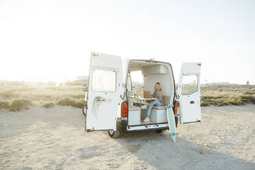 Young freelancer sitting with laptop in caravan in front of clear sky - RCPF01496