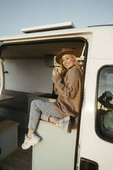 Smiling young woman with coffee cup sitting inside motor home - RCPF01525