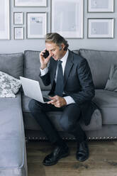 Mature businessman sitting with laptop and talking through smart phone at home - EGHF00649