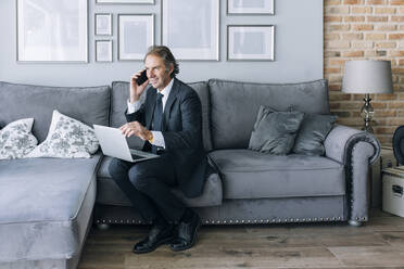 Smiling mature businessman talking through mobile phone sitting on sofa at home - EGHF00650