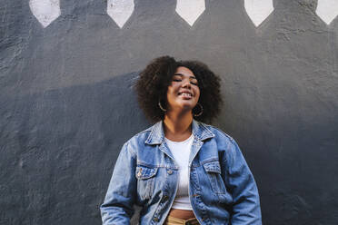 Smiling young woman with curly hair in front of wall - ASGF03103