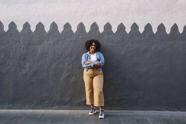 Happy woman with arms crossed standing in front of wall - ASGF03105