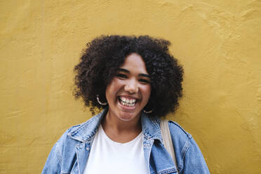 Happy young woman with curly hair in front of yellow wall - ASGF03108