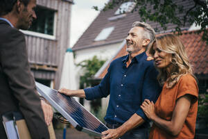 Ein glückliches Paar hält ein Solarmodul und diskutiert mit einem Immobilienmakler im Hinterhof - JOSEF14891