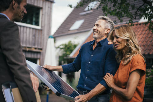 Happy couple holding solar panel and discussing with real estate agent in back yard - JOSEF14891