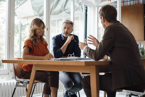 Smiling mature couple discussing over documents with real estate agent at table - JOSEF14926