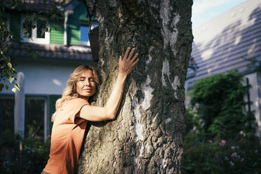 Smiling woman hugging tree in back yard on sunny day - JOSEF14951