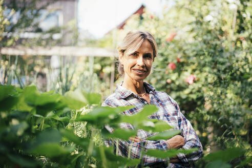 Selbstbewusste reife Frau mit verschränkten Armen inmitten von Pflanzen im Garten stehend - JOSEF14957