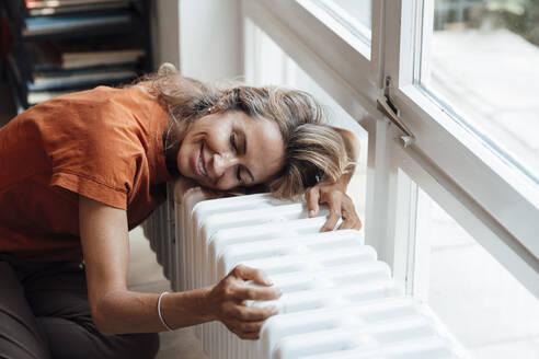 Happy mature woman leaning on radiator at home - JOSEF14970