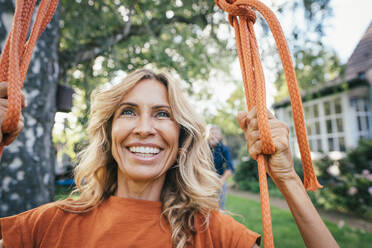 Happy mature woman sitting on swing in back yard - JOSEF14995