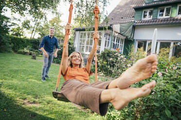 Happy woman swinging on swing with man standing behind in back yard - JOSEF14998