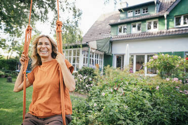 Cheerful mature woman sitting on swing in front of house - JOSEF15002