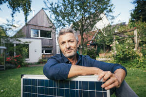 Lächelnder reifer Mann sitzt mit Sonnenkollektor vor einem Haus - JOSEF15003