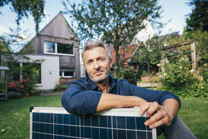 Smiling mature man sitting with solar panel in front of house - JOSEF15003