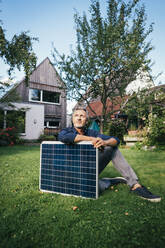 Nachdenklicher reifer Mann sitzt mit Sonnenkollektor auf dem Rasen im Hinterhof - JOSEF15004