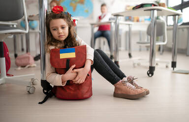 A little sad Ukrainian girl sitting on floor at school, concept of enrolling Ukrainian kids to schools. - HPIF00006