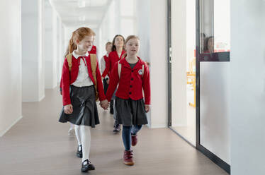 Ein glückliches Schulmädchen mit Down-Syndrom in Uniform, das sich mit einem Klassenkameraden an den Händen hält, geht in einem Schulkorridor mit Mitschülern hinter ihnen. - HPIF00189