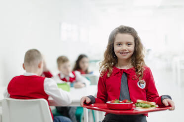 Ein glückliches Schulmädchen in Uniform hält ein Tablett mit Mittagessen in der Schulkantine. - HPIF00194