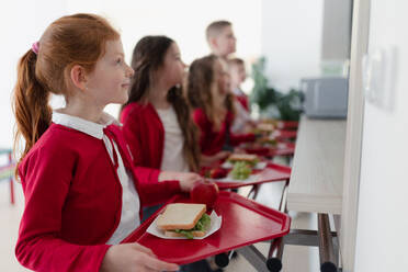Glückliche Schulkinder stehen mit Tabletts in einer Schlange und erhalten ihr Mittagessen in der Schulkantine. - HPIF00197