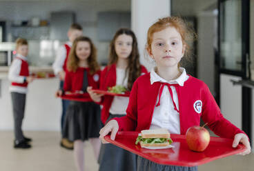 Fröhliche Schulkinder in Uniformen, die Tabletts mit Mittagessen in der Hand halten und in einer Schlange in der Schulkantine stehen. - HPIF00199