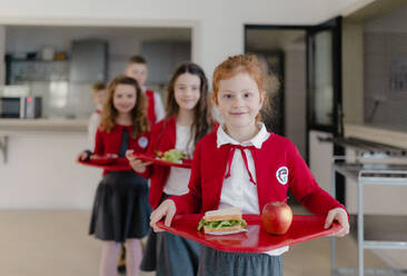 Fröhliche Schulkinder in Uniformen, die Tabletts mit Mittagessen in der Hand halten und in einer Schlange in der Schulkantine stehen. - HPIF00200