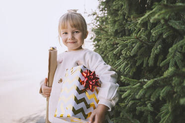 Smiling blond girl with gift box and wrapping paper near fir tree - NDEF00222