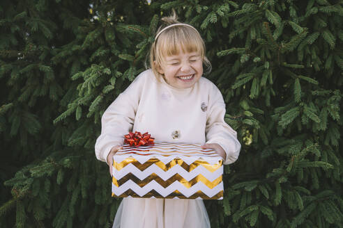 Aufgeregtes Mädchen mit einer Geschenkbox vor einem Tannenbaum - NDEF00223