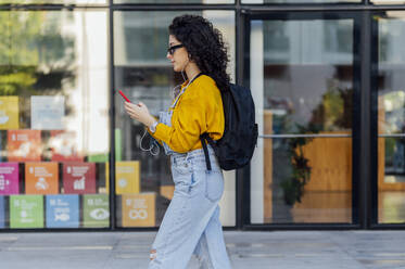 Woman listening to music through in-ear headphones and using smart phone in front of glass building - PGF01387