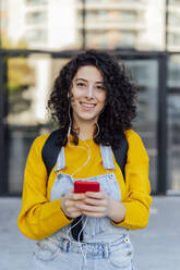 Smiling woman with mobile phone listening to music through in-ear headphones - PGF01389