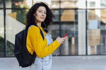 Schöne Frau mit Rucksack, die ihr Handy in der Hand hält und über In-Ear-Kopfhörer Musik hört, auf einem Fußweg - PGF01394