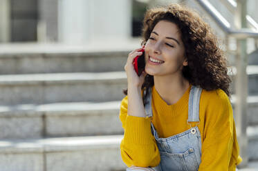 Lächelnde junge Frau mit lockigem Haar, die auf einer Treppe über ein Mobiltelefon spricht - PGF01407