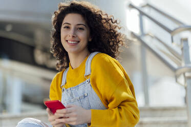 Smiling woman with curly hair holding mobile phone - PGF01410