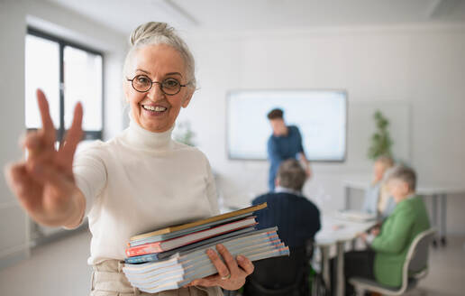 A happy senior woman student with books raising hand and looking at camera in classroom. - HPIF00420