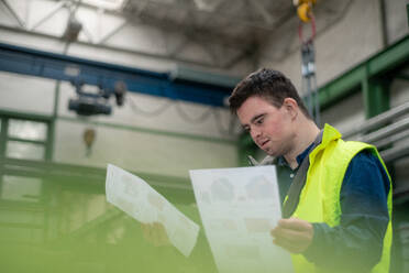 A young man with Down syndrome looking at blueprints when working in industrial factory, social integration concept. - HPIF00515