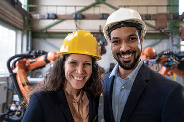 Ein Chefingenieur und Projektleiter in einer modernen Industriefabrik mit Roboterarmen schaut in die Kamera. - HPIF00582