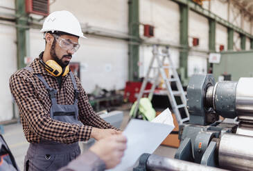 Ein Schwerindustriearbeiter mit Sicherheitskopfhörern und Schutzhelm in einer industriellen Fabrik, der Blaupausen hält - HPIF00628