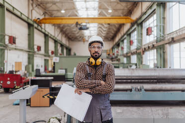 Ein Schwerindustriearbeiter mit Sicherheitskopfhörern und Schutzhelm in einer industriellen Fabrik, der Blaupausen hält - HPIF00631