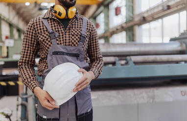 A midsection of heavy industry worker with safety headphones and hard hat in industrial factory - HPIF00633