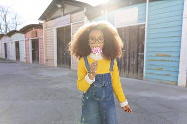 Girl eating lollipop standing near beach huts on sunny day - MEGF00198