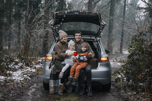 Glückliche Familie mit Kaffeetassen, die gemeinsam Zeit im Kofferraum eines Autos verbringen - NDEF00241