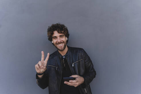 Happy man holding mobile phone gesturing peace sign leaning on gray wall - FMOF01573