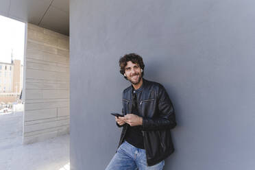 Happy man holding mobile phone leaning on gray wall - FMOF01574