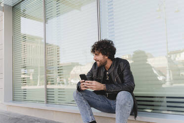 Smiling man holding mobile phone sitting on window sill - FMOF01576