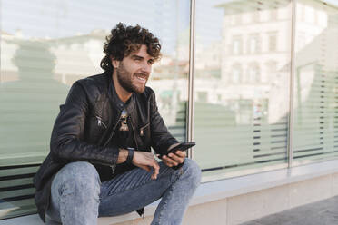 Smiling man holding smart phone sitting on window sill - FMOF01577