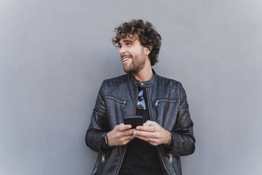 Glücklicher Mann in schwarzer Lederjacke mit Handy vor einer grauen Wand - FMOF01581