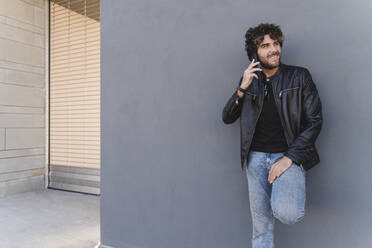 Lächelnder Mann, der vor einer grauen Wand mit einem Mobiltelefon spricht - FMOF01582