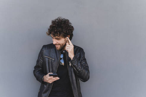Mann hört Musik über drahtlose In-Ear-Kopfhörer und hält sein Mobiltelefon an eine graue Wand gelehnt - FMOF01584