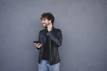 Glücklicher Mann mit Mobiltelefon, der drahtlose In-Ear-Kopfhörer vor einer grauen Wand einstellt - FMOF01586