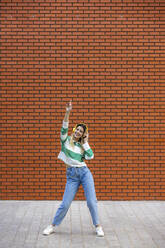 Fröhliche junge Frau, die über Kopfhörer Musik hört und auf dem Gehweg vor einer Backsteinmauer tanzt - WPEF06693