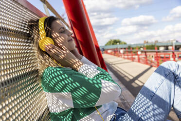 Smiling young woman listening to music through headphones leaning on fence - WPEF06705